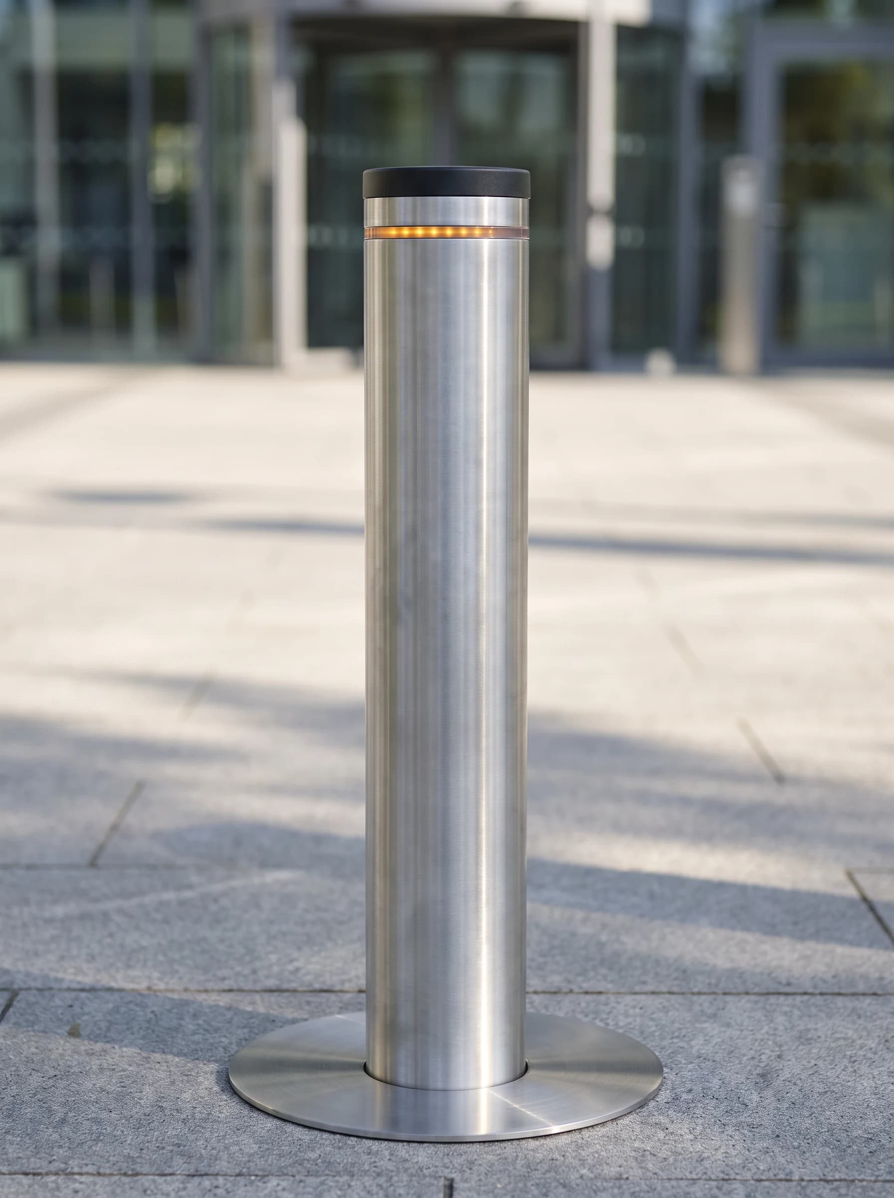 Close-up of one raised rising bollard: a brushed stainless cylinder with a flat black cap and a single amber LED band beneath it, standing in its flush stainless ground collar on stone paving