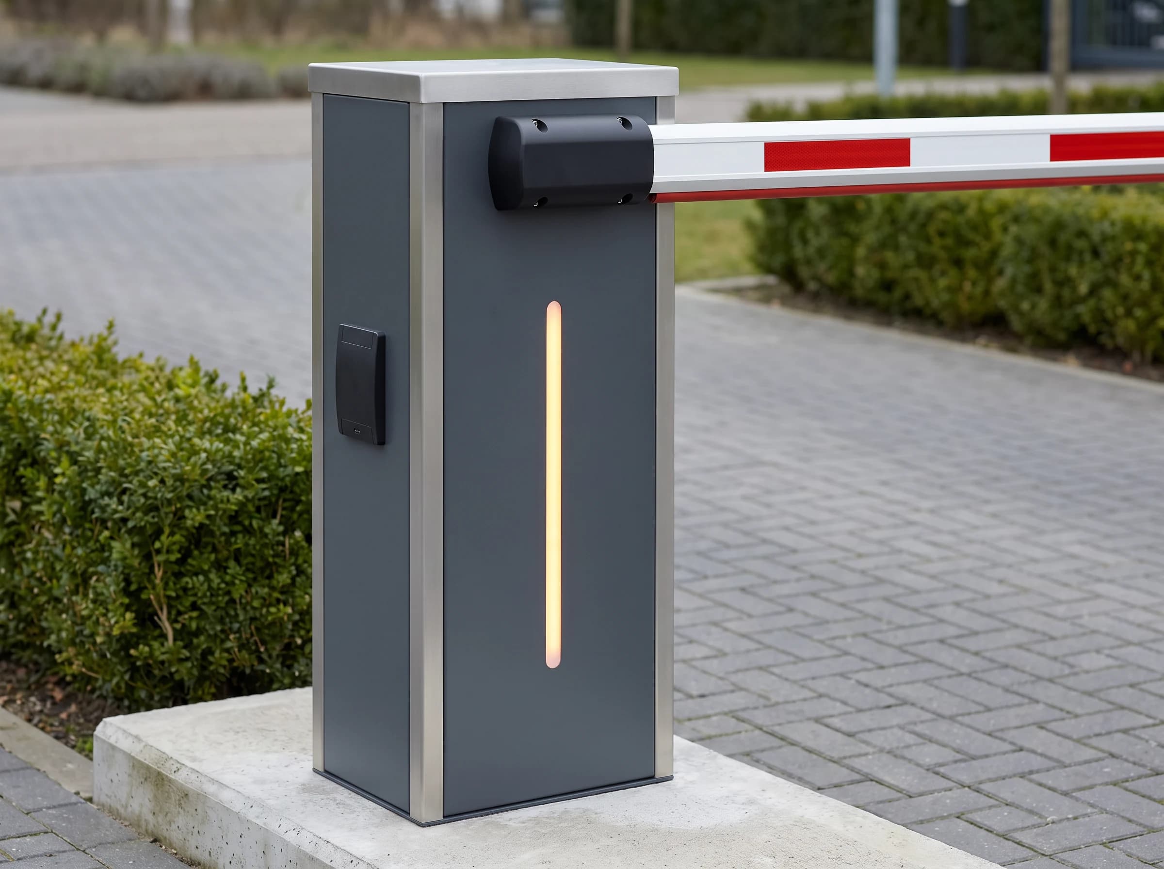 Close-up of a boom barrier operator cabinet: graphite-grey front face with brushed silver side panels and top cap, a vertical amber status strip, a card reader plate on the side and the black boom boss with the lowered white and red arm leading out of frame