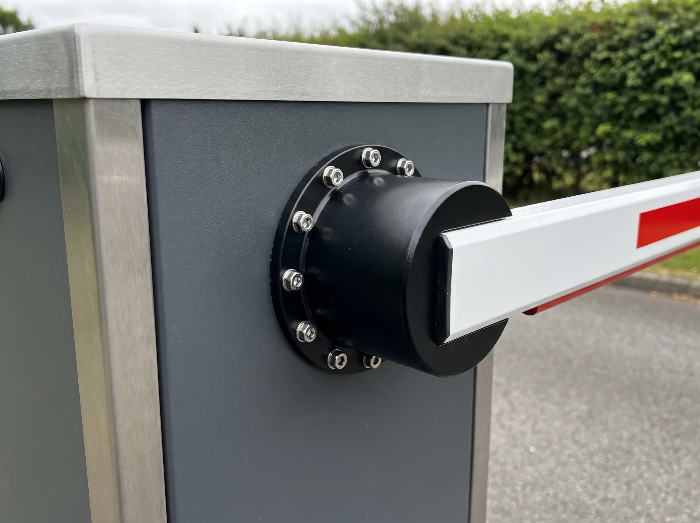 Detail of a boom barrier's boom boss: a black cylindrical boss with a ring of stainless bolts fixed to the graphite-grey side face of the cabinet below the brushed silver top cap, with the root of the white arm and its first red reflective band clamped into it