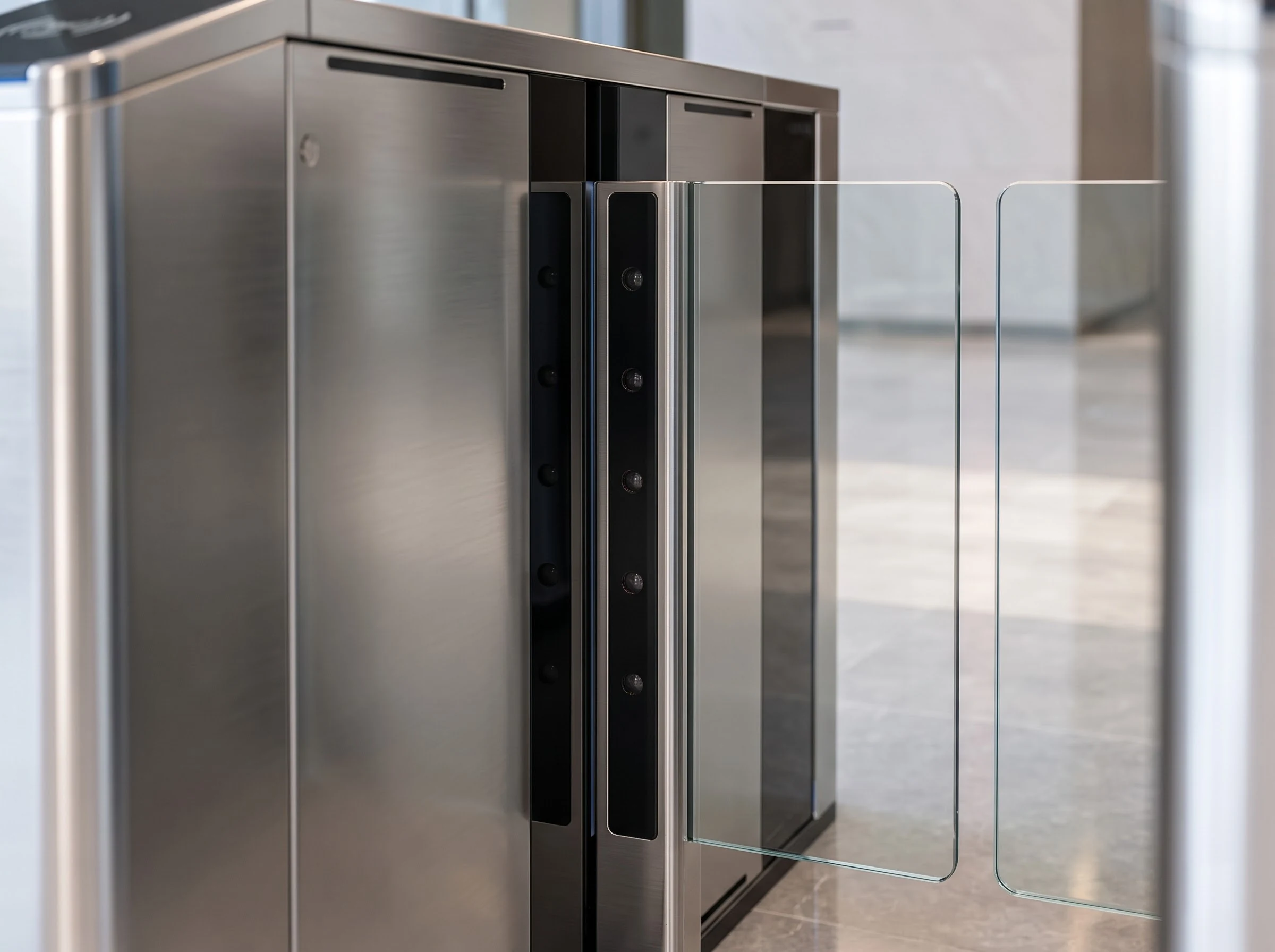 Looking along the inside of a flap barrier lane: the black glass sensor panels on the inner faces of the housings with rows of small infrared sensor windows at different heights, the glass wings at the end of the lane and the polished floor