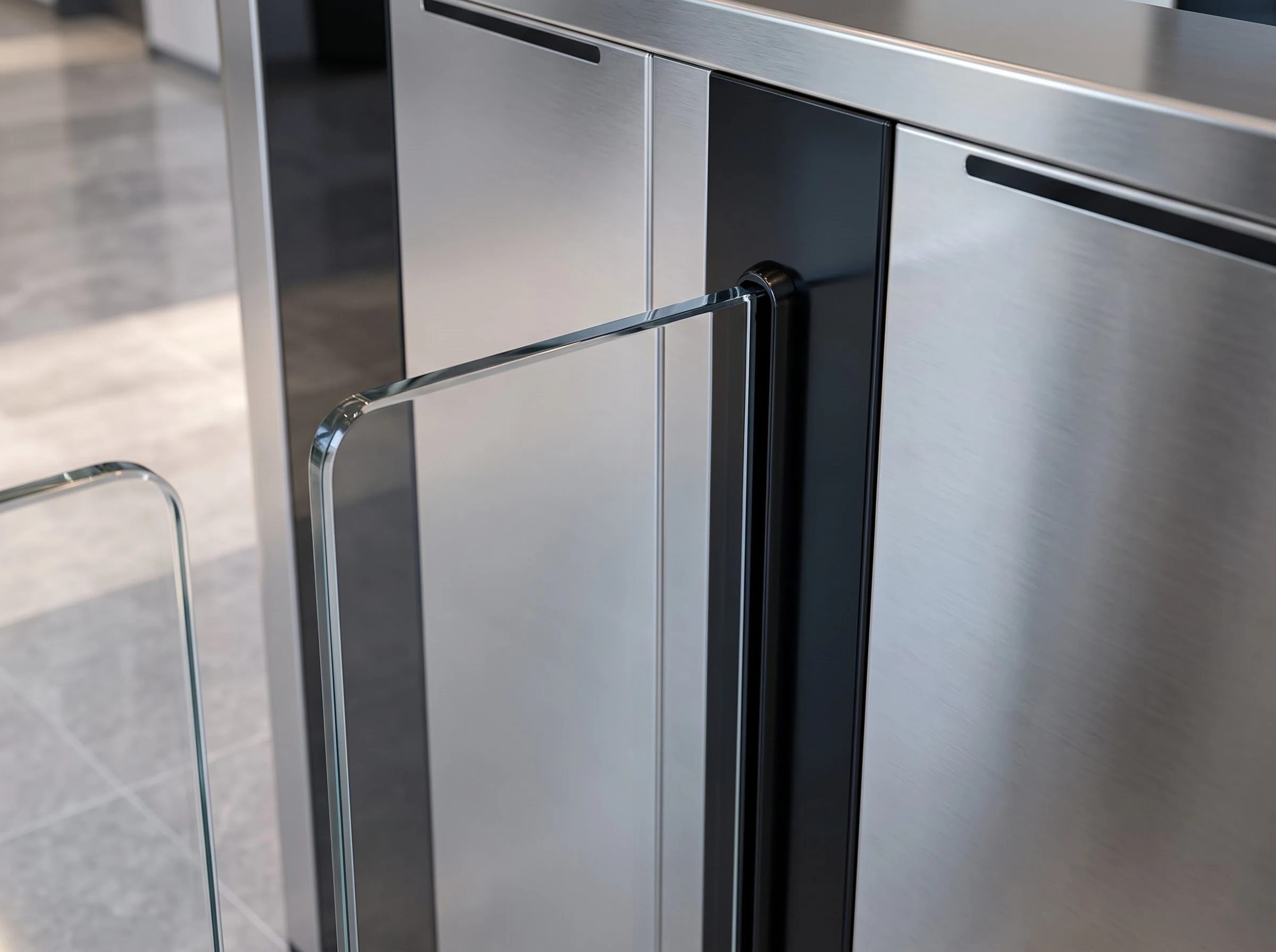 Close-up of a toughened glass wing where it emerges from the black-lined slot in the brushed stainless housing of a flap barrier, the polished glass edge reflecting the lobby