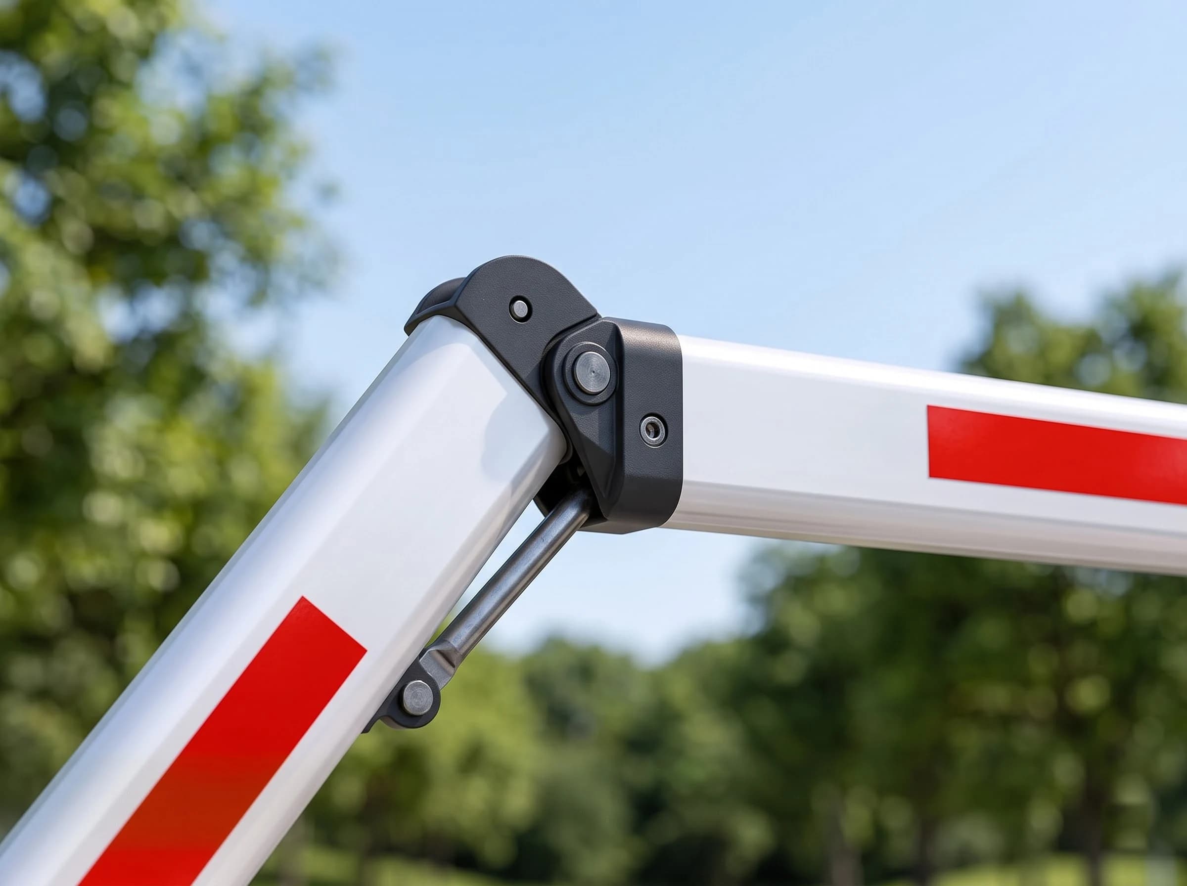 Close-up of the articulation joint of a folding-arm boom barrier: the black knuckle hinge between the two white aluminium boom sections with its pivot and the link rod that folds the outer section as the boom rises, red reflective bands on each side against trees and sky