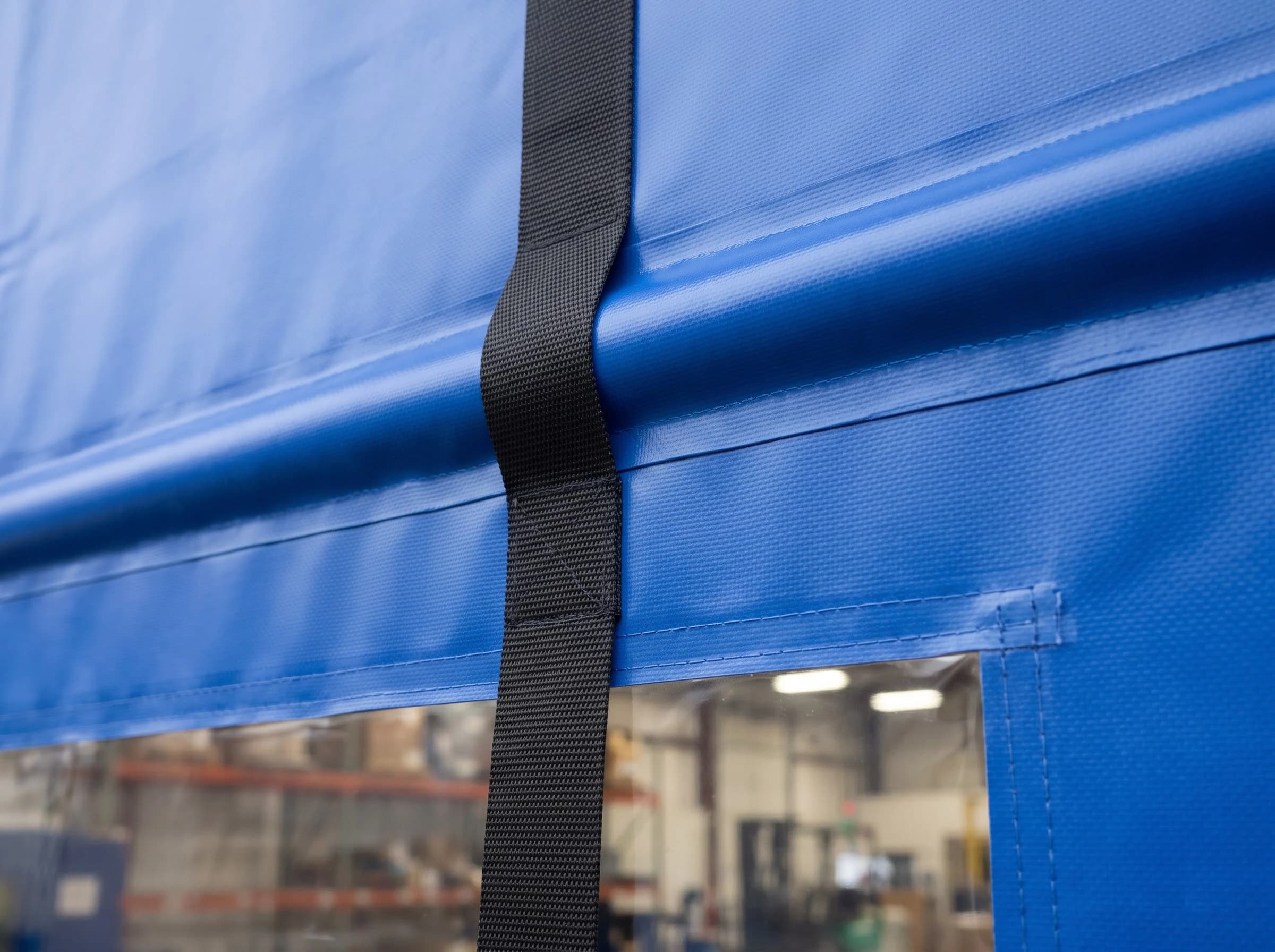 Close-up of the blue reinforced PVC curtain of a high speed fold-up door: the rounded ridge of a wind-bar pocket, a black woven lifting strap crossing it, the welded edge of a clear vision window and the fabric's woven scrim texture