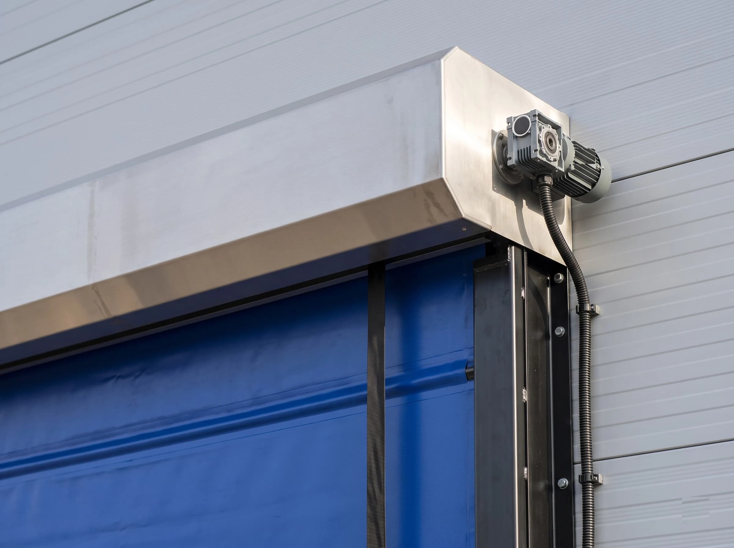 Top corner of a high speed fold-up door: the plain stainless hood with the geared electric drive motor on the strap drum shaft and its conduit, the blue curtain with wind-bar ridges and a lifting strap running up into the hood beside the black guide column