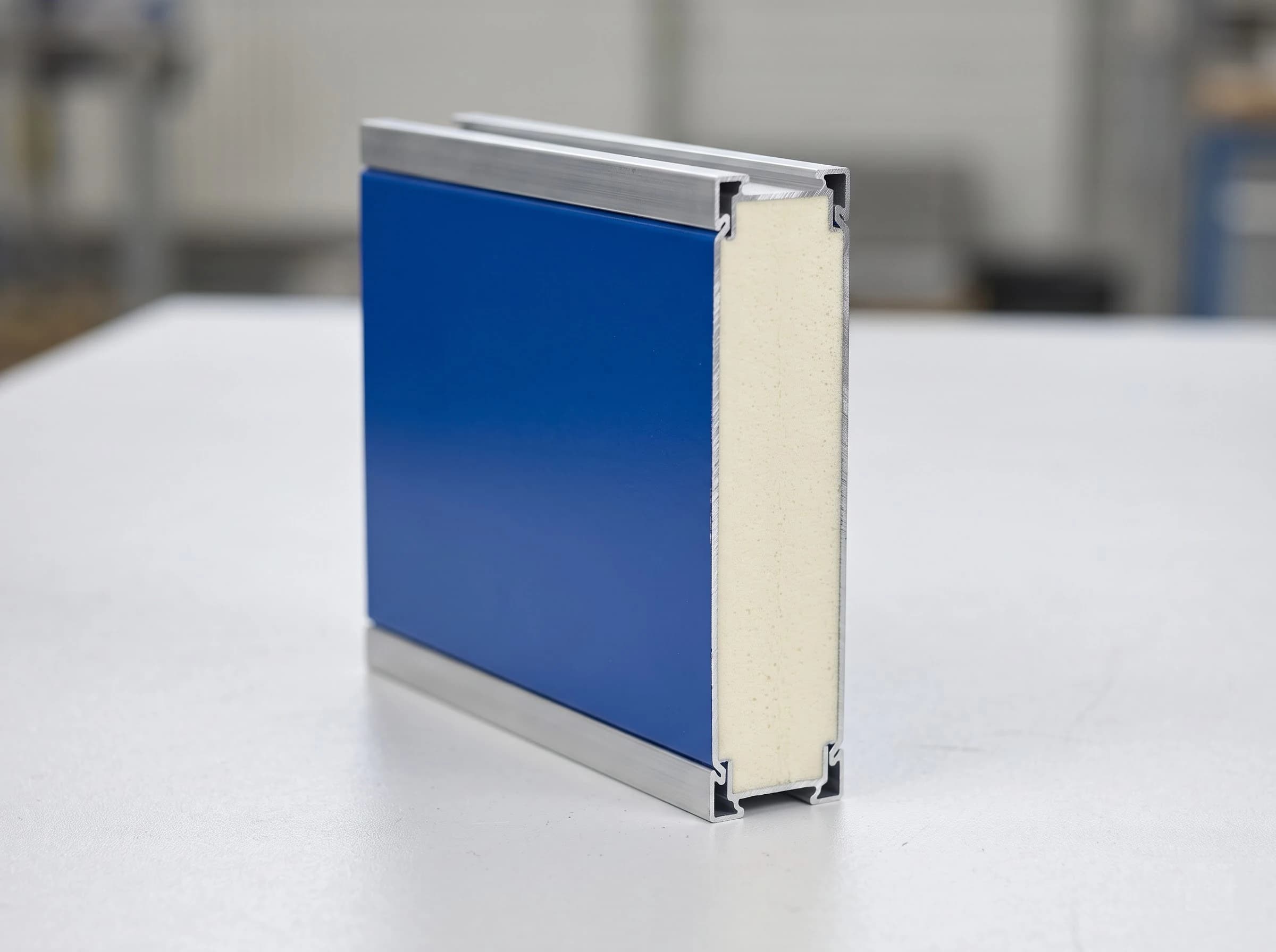 A cut sample of a high speed rigid insulated door panel on a bench: blue metal outer and inner skins with the rigid polyurethane foam insulation core filling the thickness between them, and aluminium joint profiles along the top and bottom edges