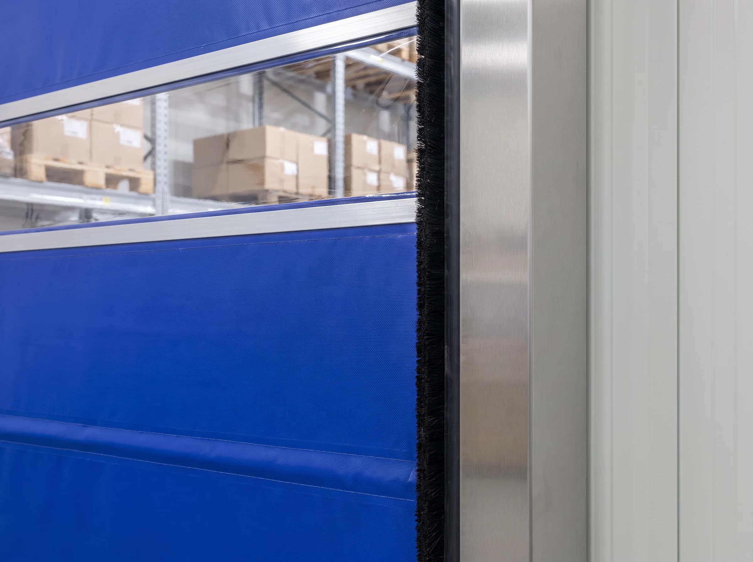 Close-up of the edge of a blue PVC high speed roll-up door: the brushed stainless guide column with its black brush seal, the curtain's reinforcing seam and the aluminium edge strip of the clear vision band