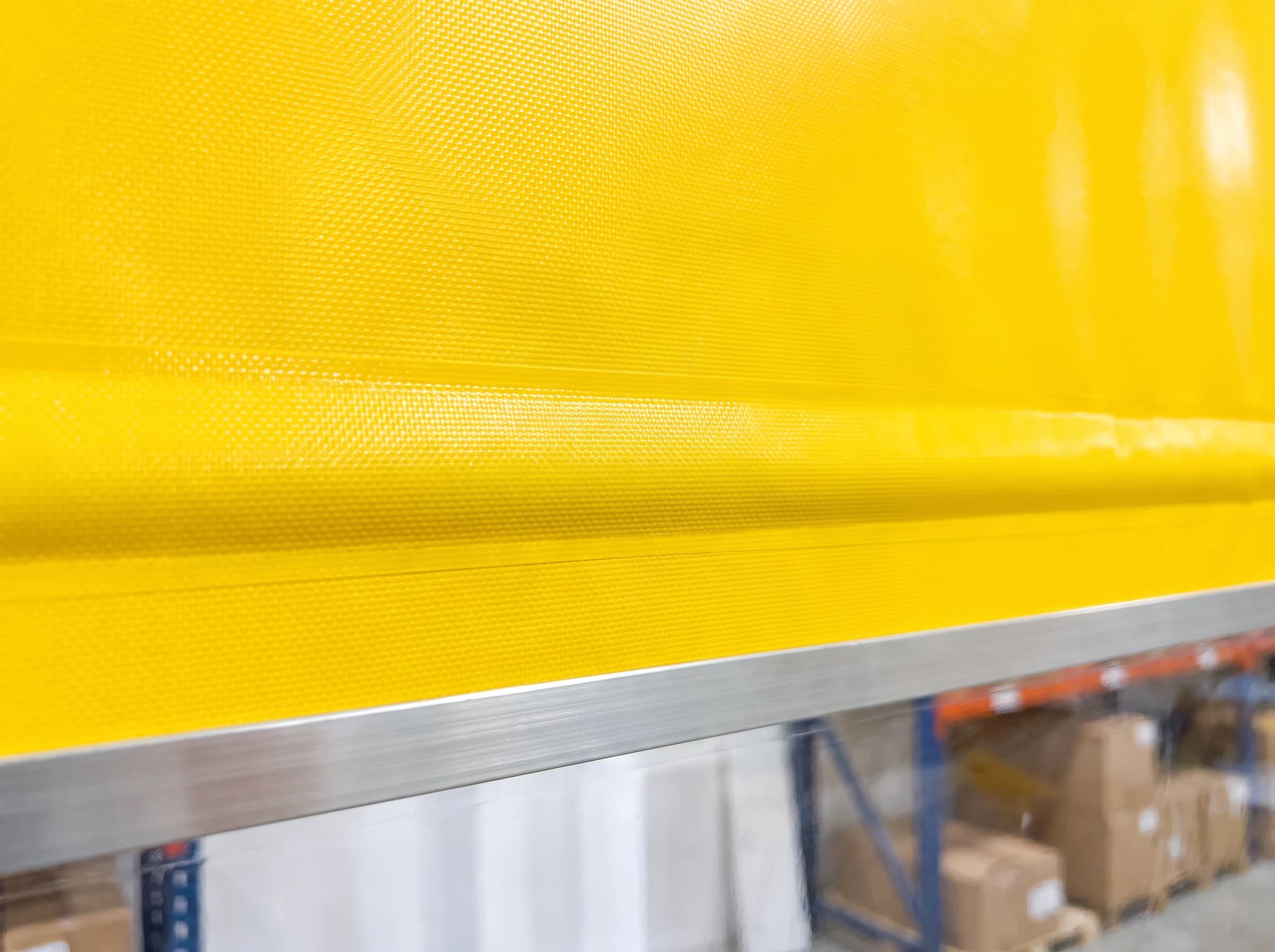 Close-up of the yellow reinforced PVC curtain of a high speed self-repairing door, showing the woven scrim texture under the coating, a welded horizontal reinforcing seam and the aluminium edge strip of the clear vision band