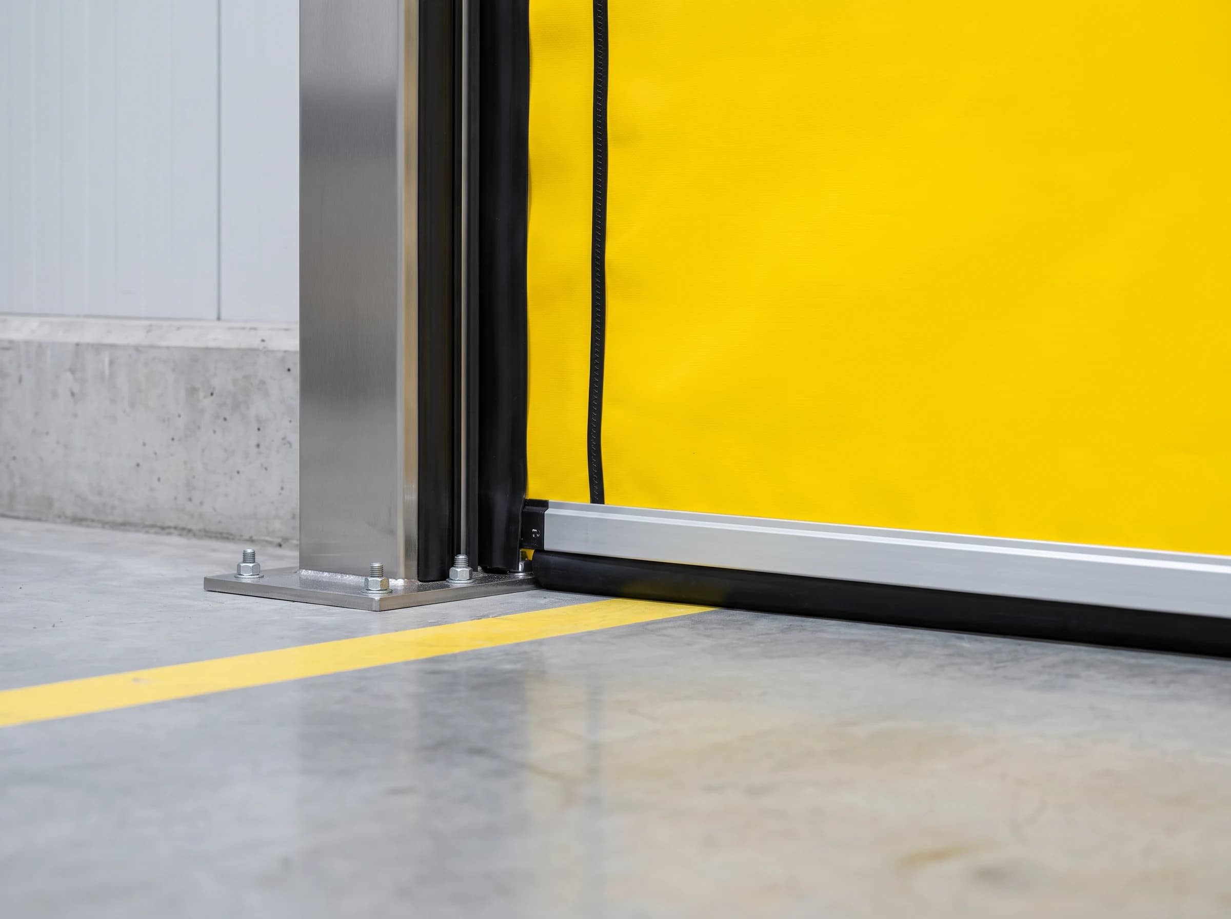 Bottom corner of a high speed self-repairing door: the aluminium bottom rail with its black soft floor seal, the foot of the stainless guide column bolted to the floor, and the curtain's edge bead running in the black flexible guide channel