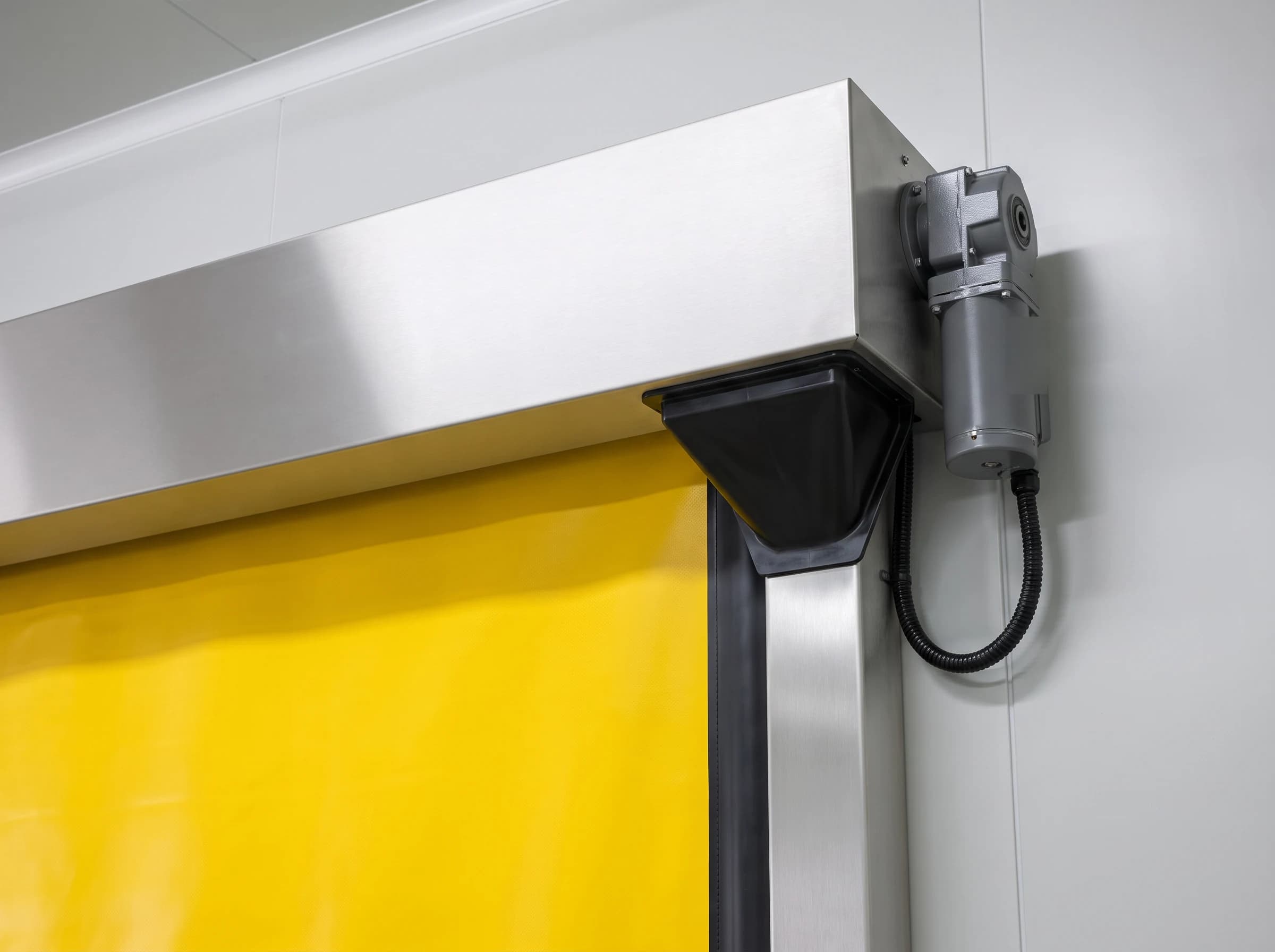 Top corner of a high speed self-repairing door: the plain stainless hood over the roller, the flared re-entry mouth at the head of the guide that re-threads the curtain edge, and the geared electric operator on the roller shaft end