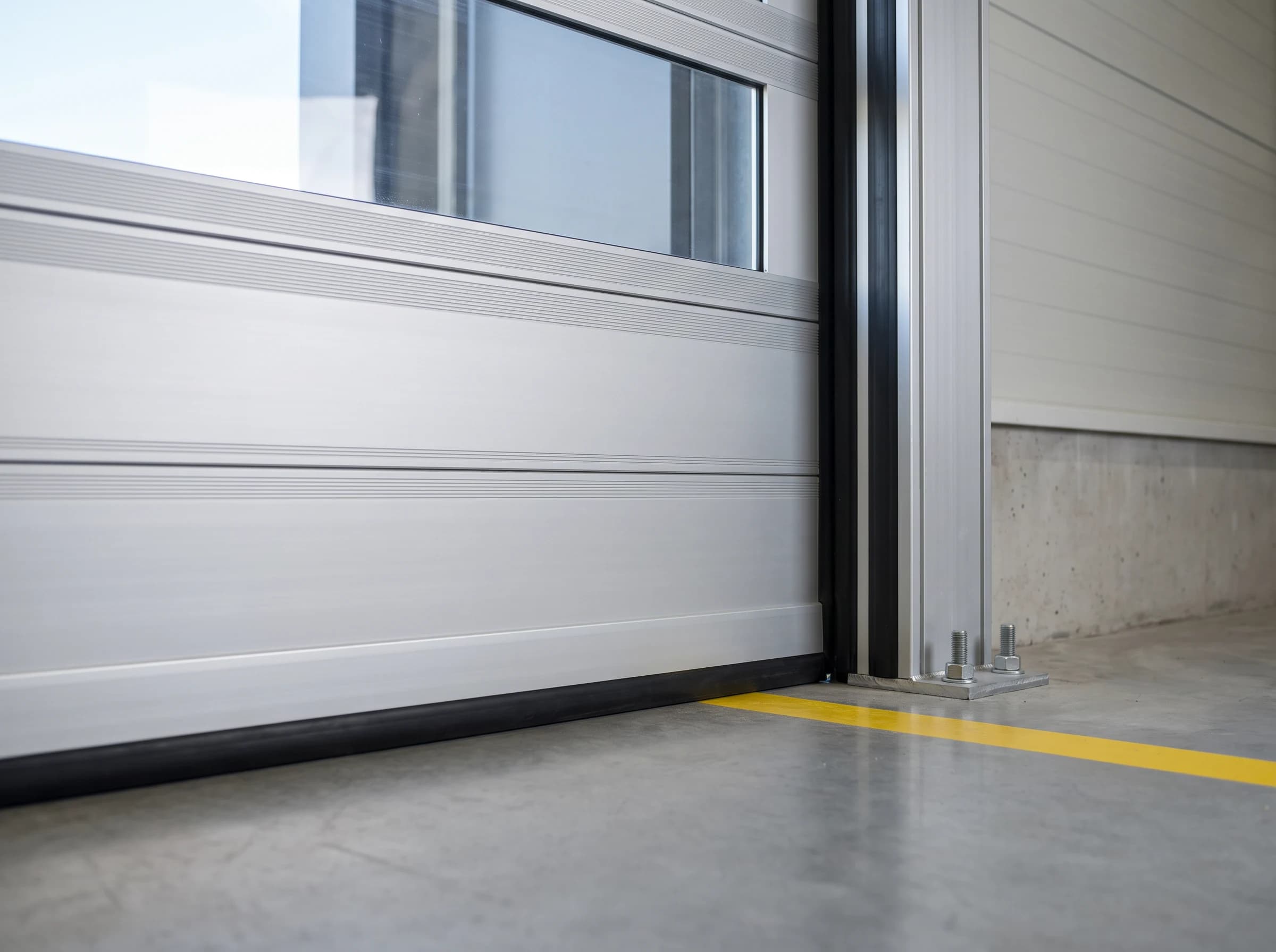 Bottom corner of a closed high speed spiral door: the leading aluminium panel with its black rubber safety edge on the concrete, a glazed panel above, and the foot of the guide column bolted to the plinth with its black seal
