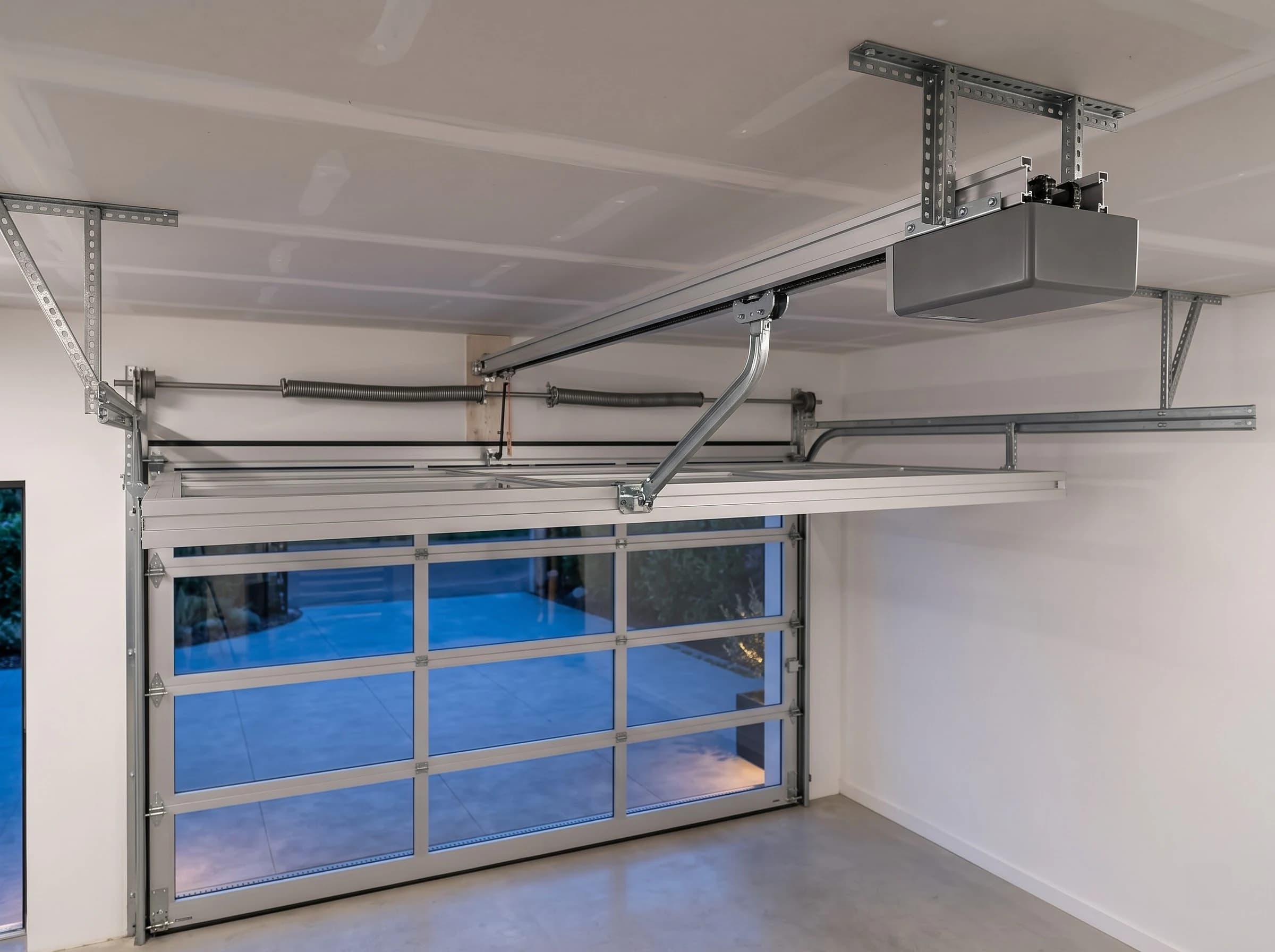 Inside view of an aluminium garage door's drive: the ceiling-mounted operator on its brackets, the drive rail and trolley arm connected to the top section, the torsion spring above the door head and the horizontal tracks, with the clear-glazed door below