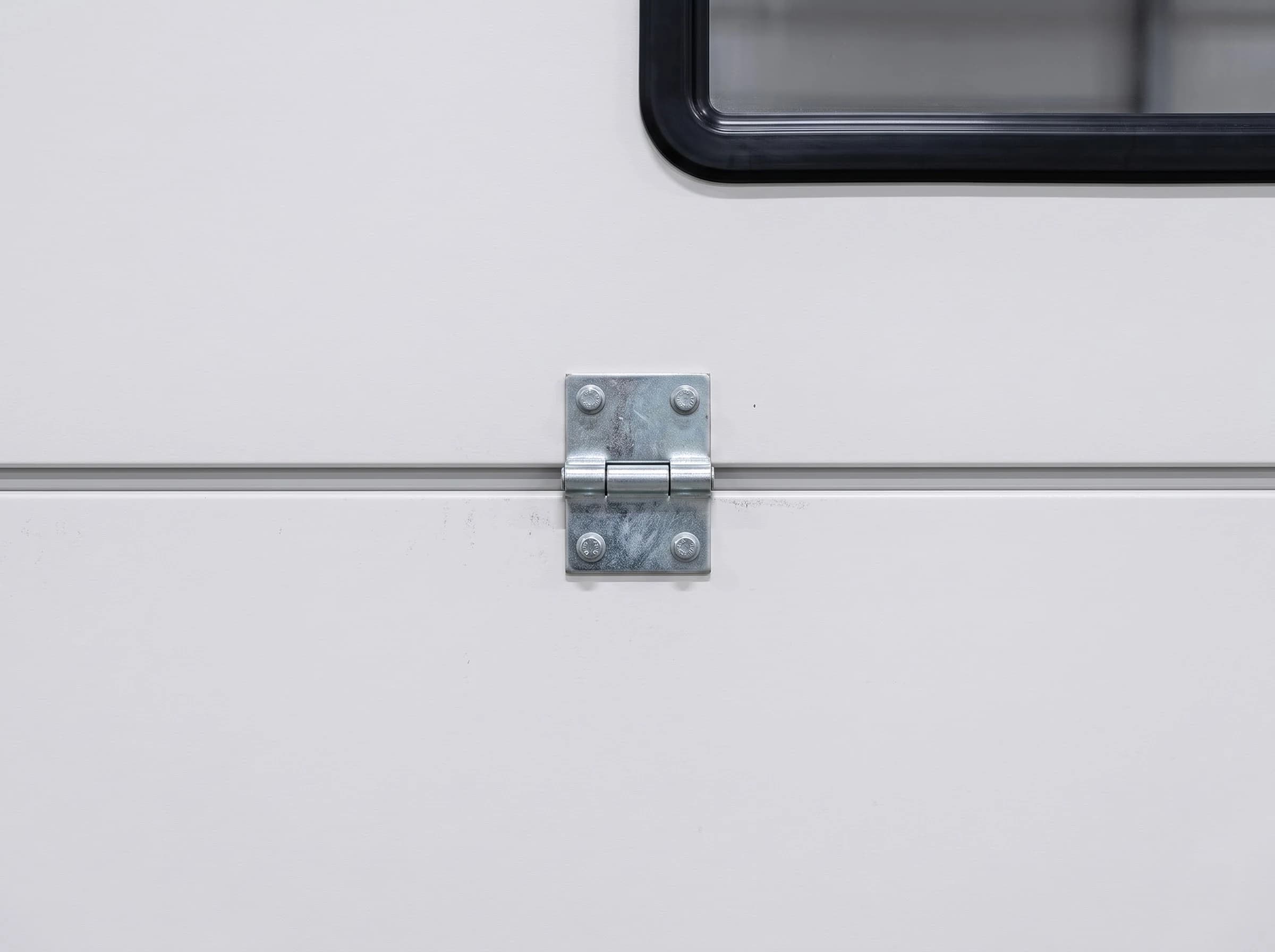 Square-on close-up of a sectional door panel joint: two flush white insulated panels meeting along a level joint line, a galvanised intermediate hinge bolted across it and the corner of a black-framed window above