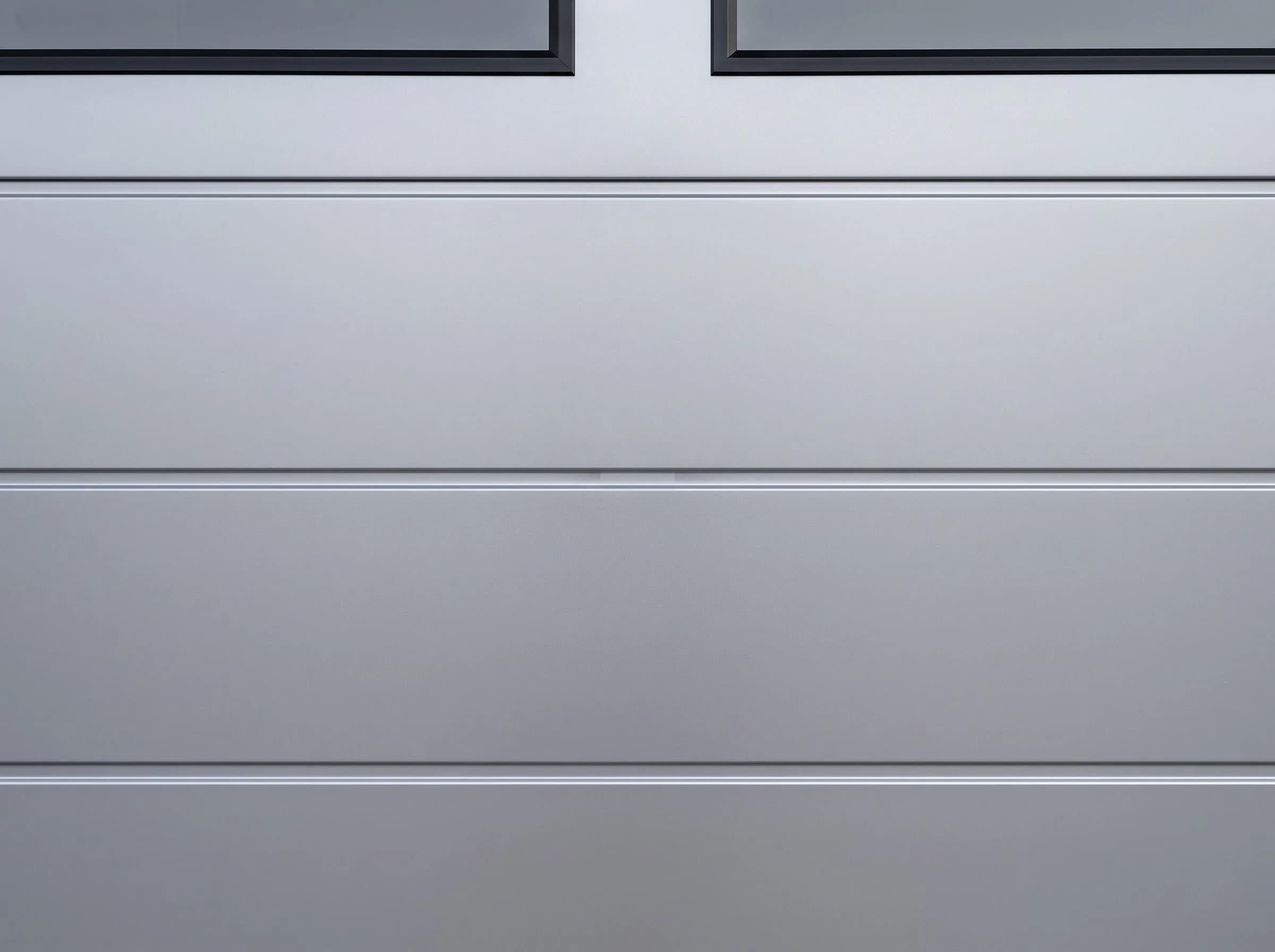 Square-on close-up of a residential garage door face: two flush silver-grey steel sections meeting along a level rounded joint, each with fine horizontal groove lines, and the lower edge of the black-framed windows above
