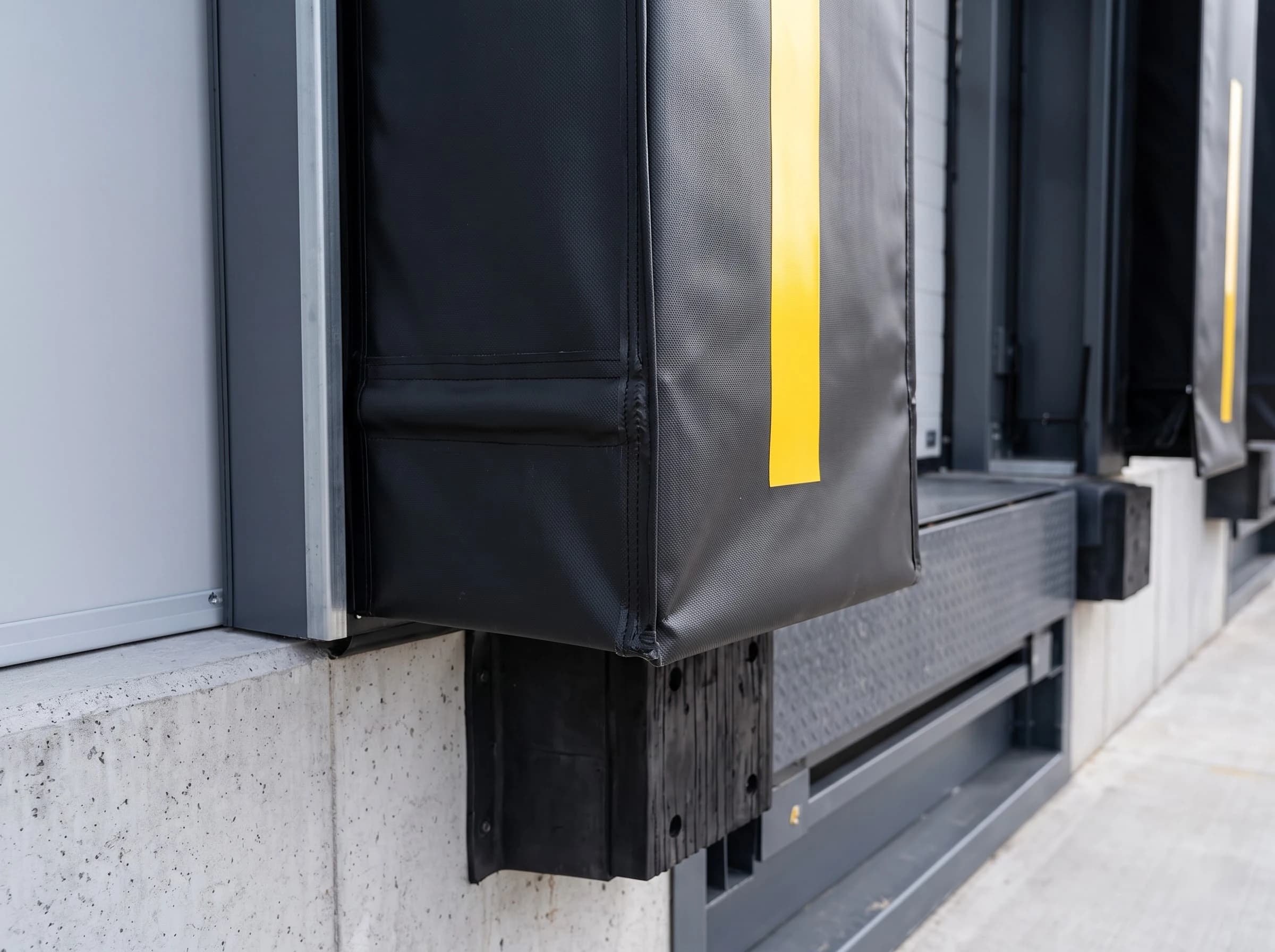 Close-up of the foot of a dock shelter side curtain: the heavy black PVC with its vertical yellow guide stripe and welded edge hanging beside the sectional door, with the black rubber dock bumper and the dock leveller edge below on the concrete dock face