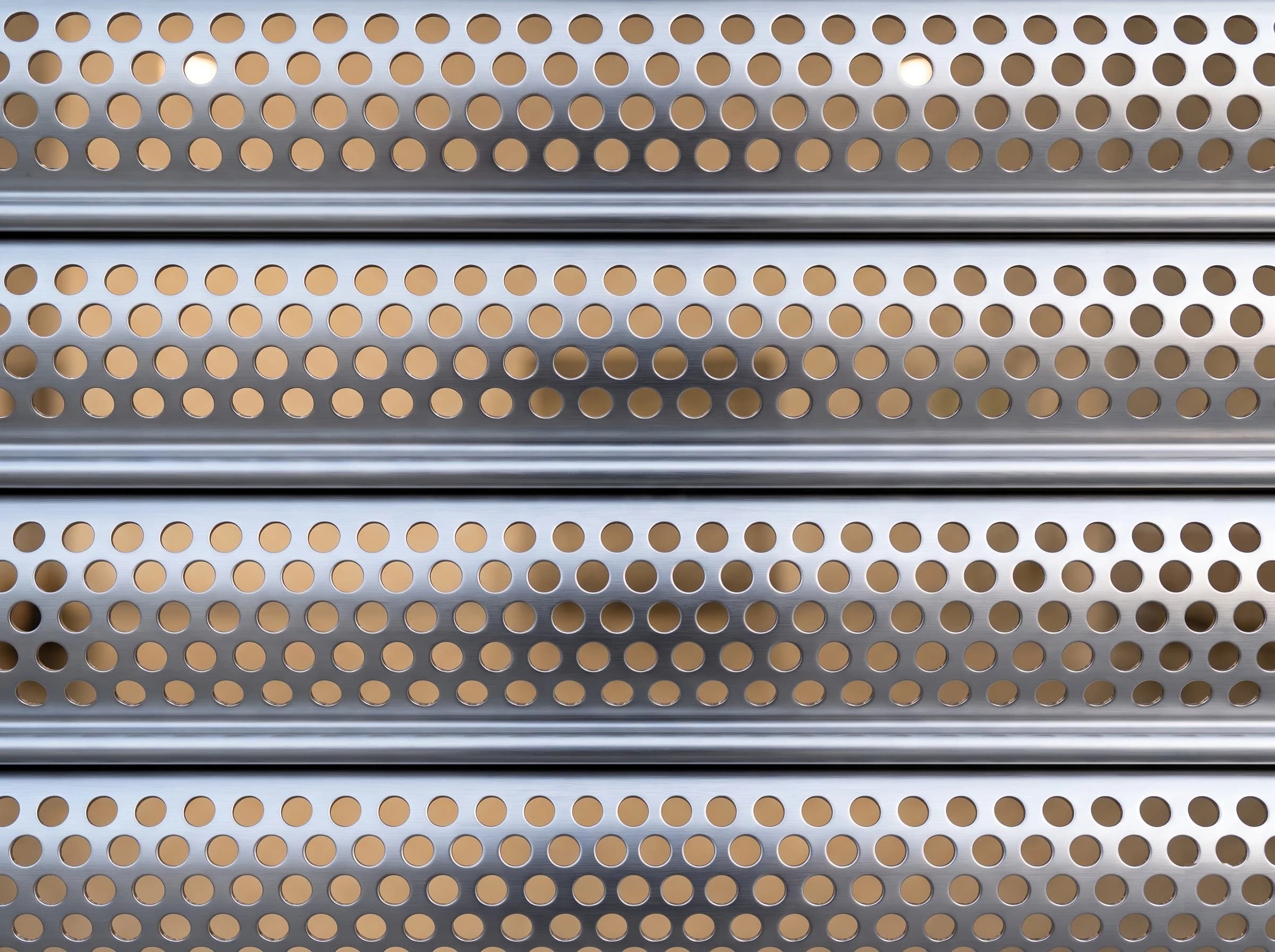 Extreme close-up of a perforated rolling shutter curtain: four curved brushed stainless slats with staggered rows of round perforations, joined by rolled interlocks, light from the showroom showing through the holes